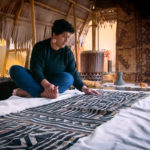 Melodie is pictured sitting inside the PIEAM fale wearing jeans and a black sweatshirt. She is looking down at the masi.